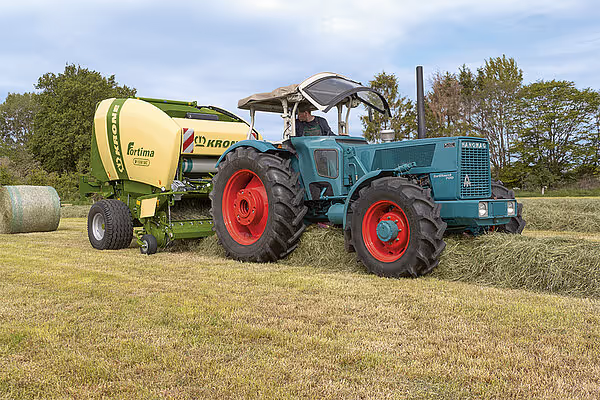 KRONE Fortima rundballpresse koblet til blå traktor med røde felger på stubbet eng. Ferdig rundball ligger ved siden av maskinen.