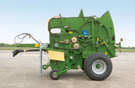 Grønn Krone Fortima rundballpresse med gul beholder, montert på hjul med trekkstang for traktor. Maskinen står på asfaltert underlag.