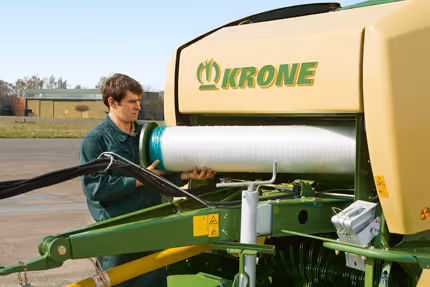 Operatør i grønn arbeidsdrakt betjener KRONE Fortima rundballpresse. Hvit plastfilm for ballinnpakning er synlig på maskinen.