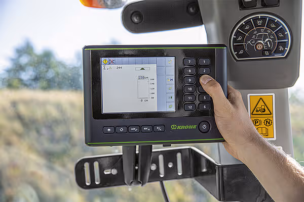 KRONE terminal med touchskjerm i traktorkabinen som viser driftsdata. En hånd opererer kontrollpanelet på det grønne displayet.