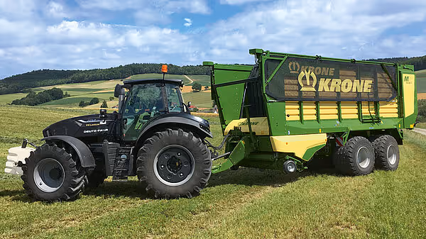 Sort traktor koblet til grønn og gul KRONE MX silohakker på et grønt slåttemark med blå himmel.