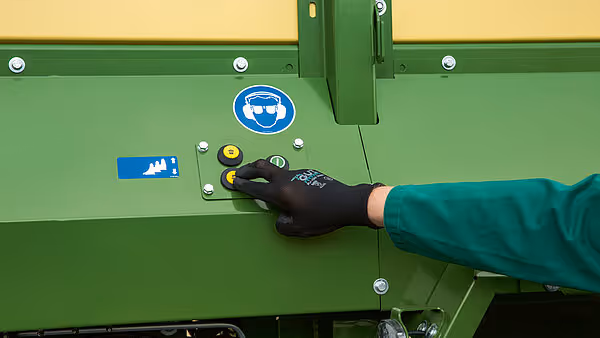 Hånd som justerer eller inspiserer sikkerhetsfunksjoner på siden av grønn KRONE landbruksmaskin.