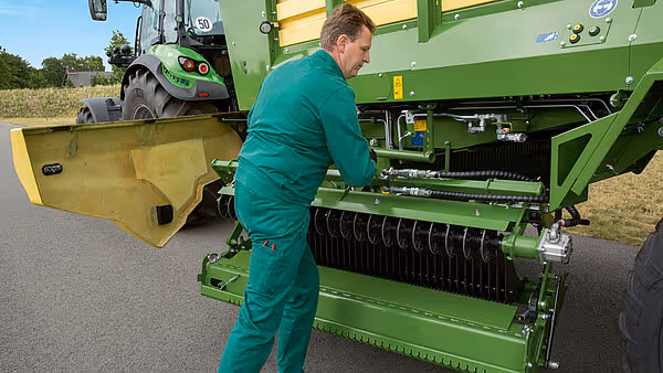 Tekniker i grønn arbeidsoverall inspiserer og justerer hydrauliske komponenter på en KRONE MX fôrvogn.