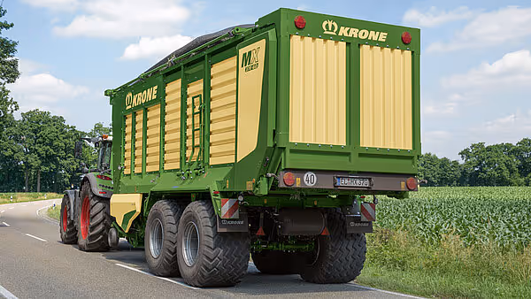 Rød traktor med påhengsvogn transporterer grønn og gul Krone MX silofyll-vogn på landevei.