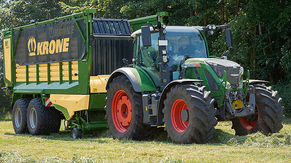 Grønn og gul KRONE MX silohakker koblet til grønn traktor med røde felger, fotografert fra siden på grønt beite med skog i bakgrunnen.
