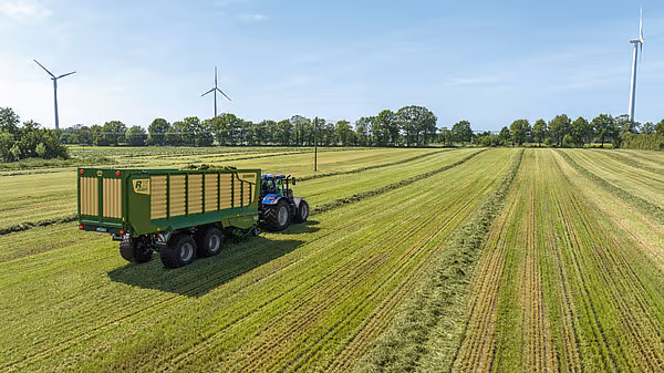 KRONE RX forvogn fyllt med rundballsilo på åker med vindmøller i bakgrunnen.