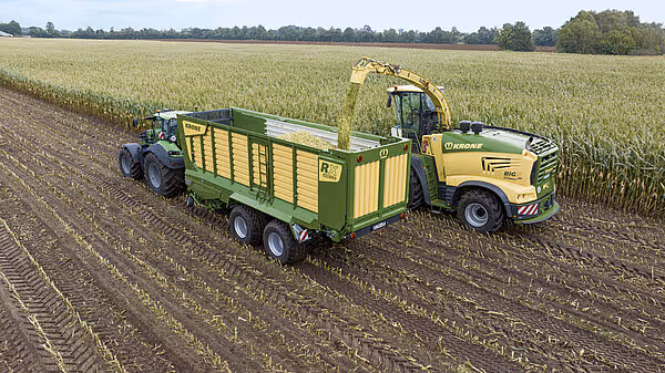 En gul maishaksleutrustning med gribarm tømmer maiss inn i en grønn og gul Krone RX vogn som trekkes av en blå traktor på et jordet.