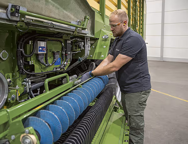 Servicemedarbeider inspiserer og justerer den blå rotorkutteraxelen på en grønn KRONE RX høstmaskin.