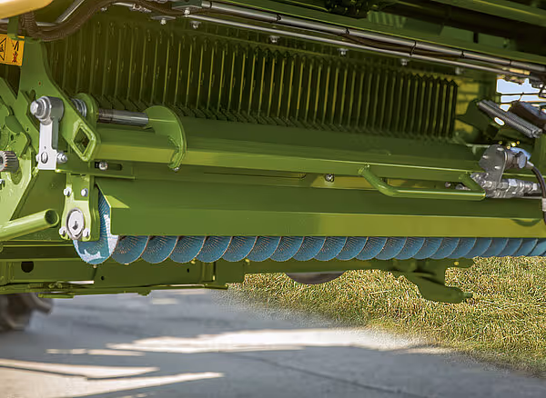 Nærbilde av hakkemaskinens skjærutstyr med blå kniver montert på grønn ramme, med høy gress synlig under maskinen.