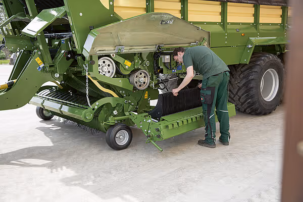 Operatør utfører service på presskammer under KRONE RX kornvogn trukket av traktor.