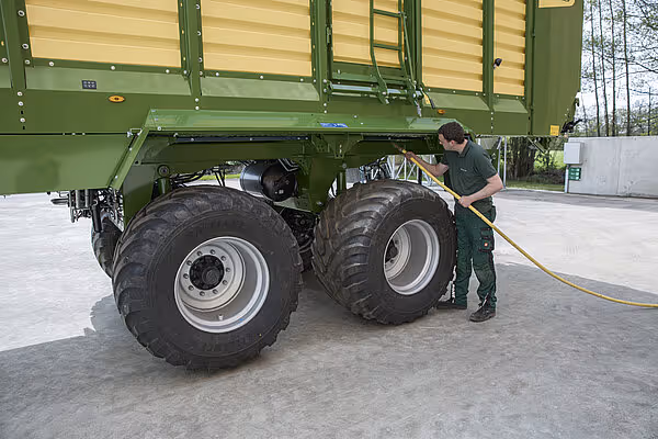 Mann vasker bakhjulet på en grønn KRONE RX vogn med høytrykksspyler på betongunterlag.