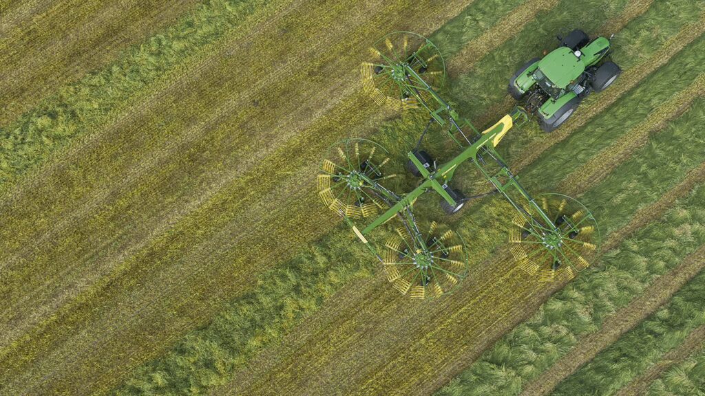 Luftfoto av KRONE Swadro TC vendehark i arbeid på eng, trukket av grønn traktor. Harken samler høy i rader med sine roterende rotorer.
