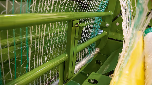 Detaljbilete av nettføringssystemet på rundballepressa. Det grøne nettet blir styrt gjennom metallskinner og rullar.