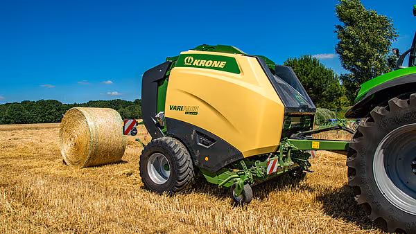 KRONE VariPack Plus rundballpresse i arbeid på stubbeåker med en ferdig rundball liggende ved siden av maskinen. Pressen er koblet til en traktor og arbeider under blå himmel.