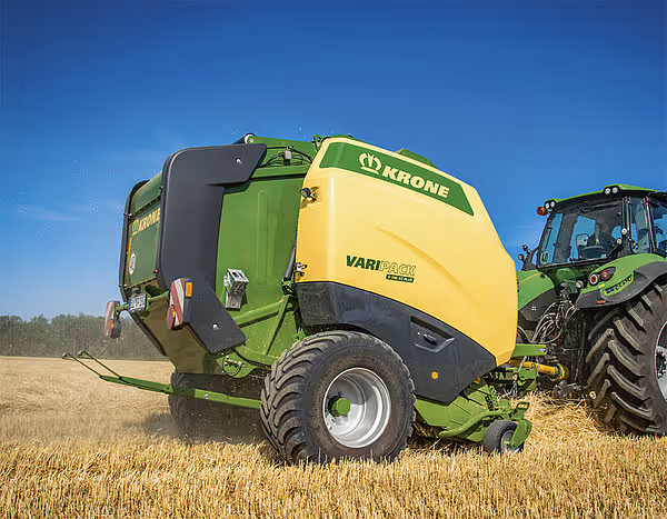 KRONE Varipack Plus rundballpresse i gul og grønn farge trukket av traktor på stubåker.