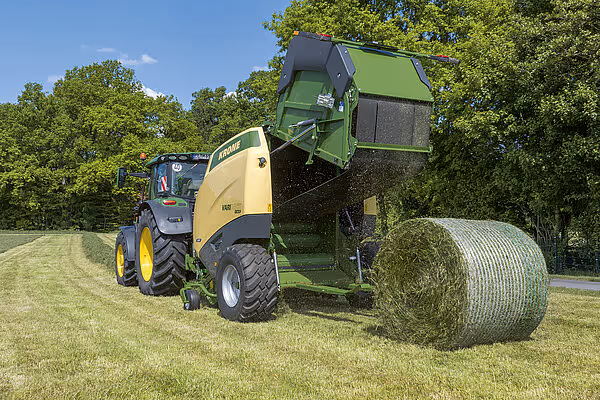 KRONE Varipack Pro rundballpresse trukket av traktor produserer stor rundball på grønt grasareal. Maskinen har grønn og gul fargekombinasjon.