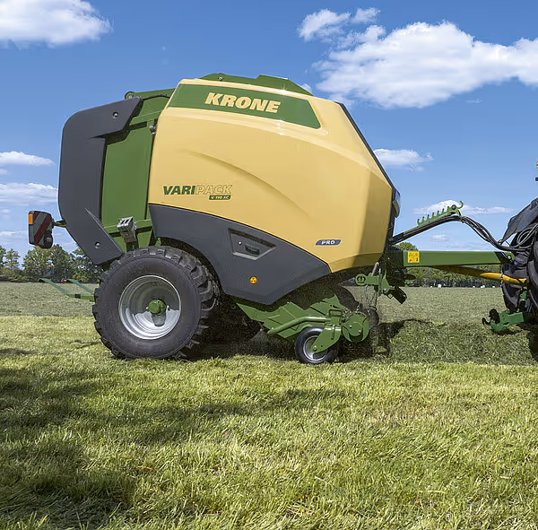 KRONE Varipack rundballpresse i arbeid på eng under blå himmel med skyer. Maskinen er tilkoblet en traktor og arbeider med grasavling.