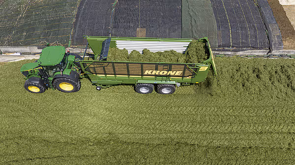 Drone-bilde av en grønn Krone ZX høstingsvogn trukket av en traktor, lastet med grønt fôr på eng.