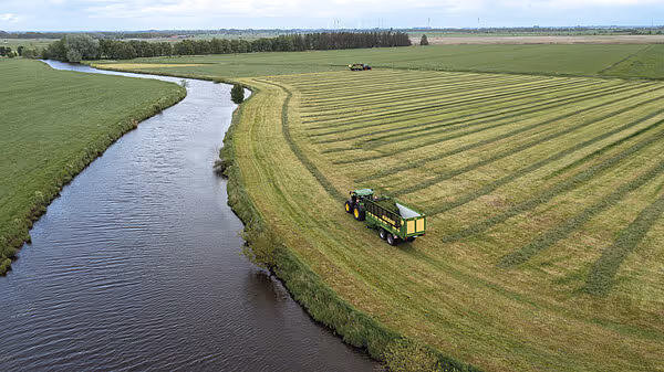 Luftfoto av traktor med Krone ZX høstingsvogn som høster gras langs en kanal i et flatt landbrukslandskap.