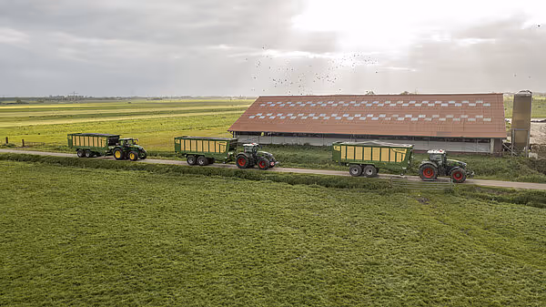 Flere traktorer med Krone ZX høstingsvogner parkert foran en stor driftsbygning ved siden av en eng.