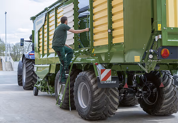 Landbruksarbeider åpner høylaster-siden på grønn KRONE ZX-vogn festet til traktor på betongflate.