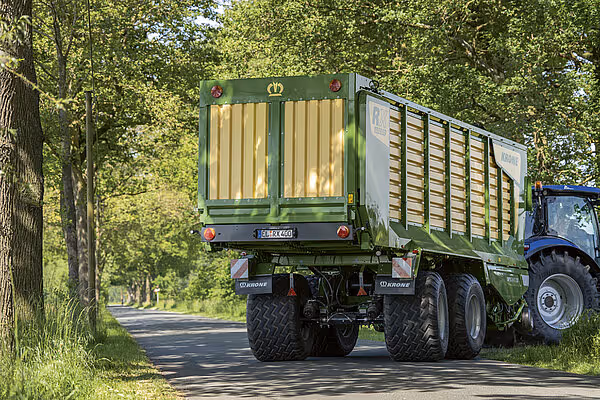 Krone ZX silofyll-vogn med grønt og gult design, trukket av blå traktor på skogsvei.