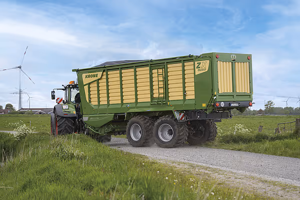 Grønn traktor med KRONE fôrvogn på hjul kjører på landbruksvei med grønt grasarealer og himmel i bakgrunnen.