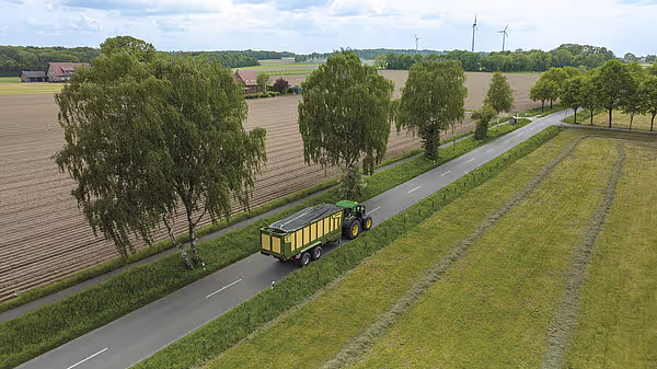 Luftfoto viser grønn traktor med gul fôrvogn som kjører på landbruksvei mellom grønne marker og treplantasje.
