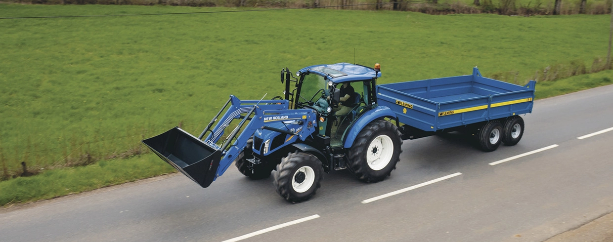 En New Holland-traktor med frontlaster kjører på veien med en blå Fleming-tilhenger bak seg.