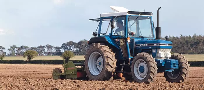 En blå Ford 4610-traktor med hvitt tak pløyer en åker under åpen himmel.