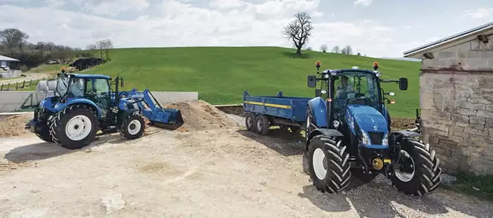 To New Holland-traktorer på en gårdsplass, en med frontlaster og en annen med en tilhenger.