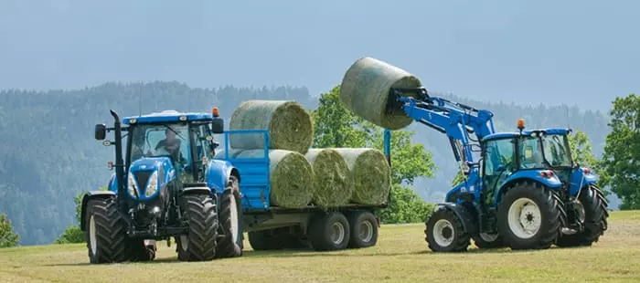 To New Holland-traktorer på en gårdsplass, en med frontlaster og en annen med en tilhenger.