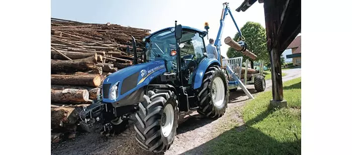 En blå New Holland-traktor i arbeid ved en tømmerplass, der den trekker en tilhenger med en hydraulisk kran som løfter tømmerstokker.