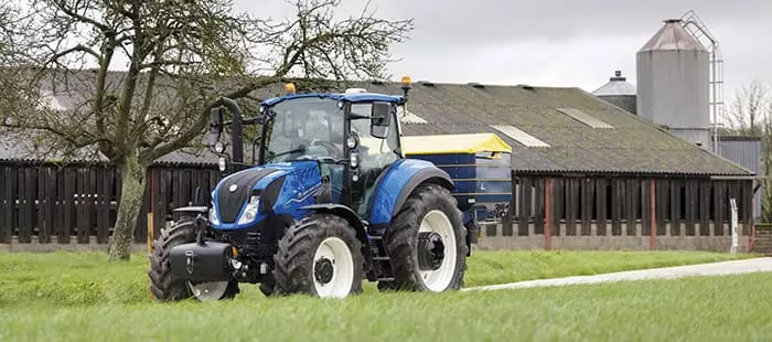 New Holland T5 traktor ved gårdsbruk – En blå New Holland T5-traktor foran en bondegård med siloer og uthus.