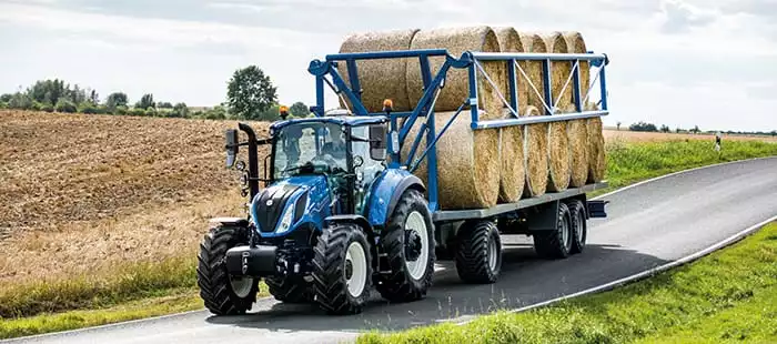 New Holland T5 transporterer høyballer – En blå New Holland-traktor trekker en tilhenger lastet med rundballer langs en landevei.