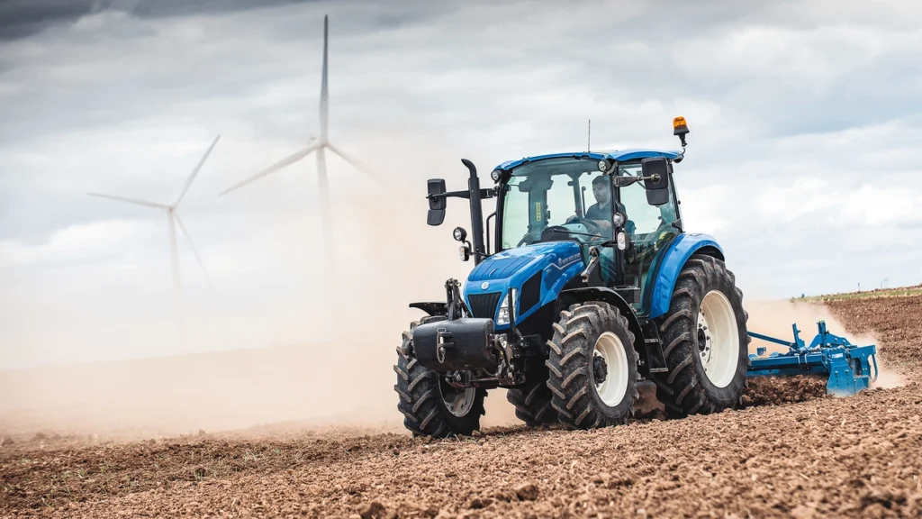 New Holland-traktor som pløyer jord, med føreren synlig i hytta. Store hjul med dype spor kaster opp støv mens traktoren beveger seg over åkeren.