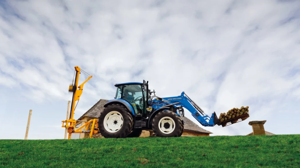 New Holland-traktor med frontlaster, som løfter en pall med tømmerstokker opp en grønn bakke. Et gult redskap er festet til baksiden.