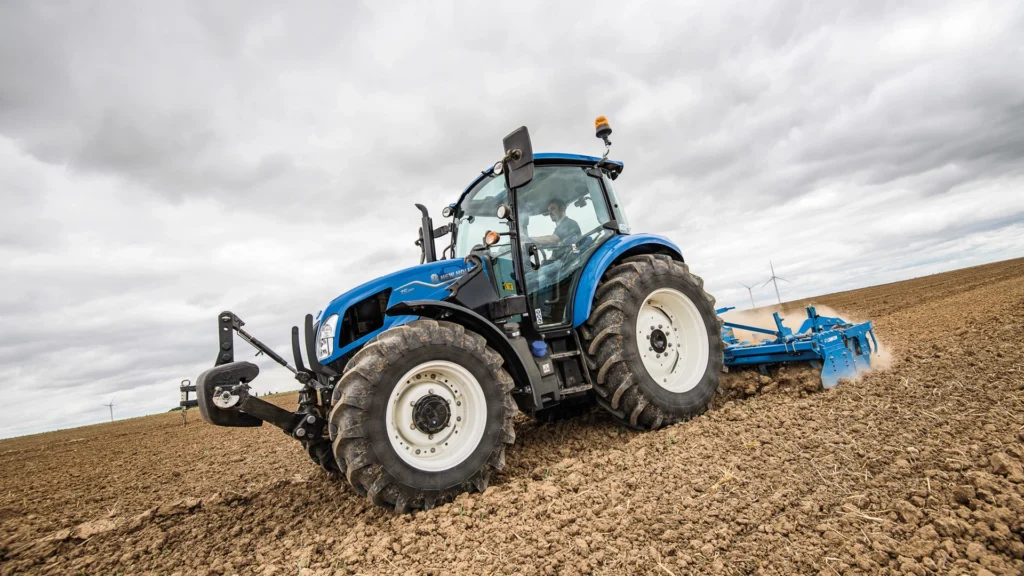 New Holland-traktor i arbeid på åker, sett fra siden, der den trekker et jordbearbeidingsredskap gjennom tørr jord. Vindmøller kan sees i bakgrunnen.