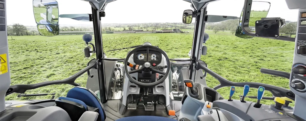 Førermiljø i en New Holland-traktor sett fra innsiden av hytta, med ratt, spaker og kontrollpanel i fokus. En åpen grønn åker vises gjennom frontruten.