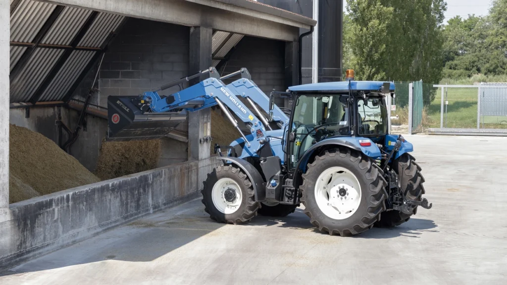 New Holland T5S-traktor med frontlaster laster fôr i en silo, med presisjonskontroll i et industrielt miljø.