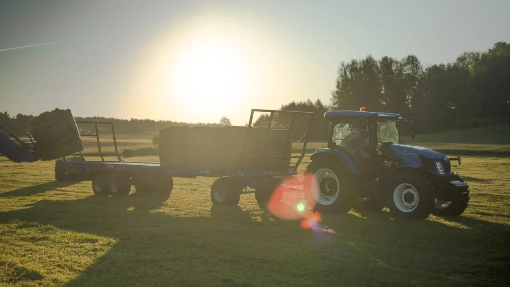 New Holland T5S-traktor trekker en henger med høyballer på en eng ved solnedgang, med varm belysning.