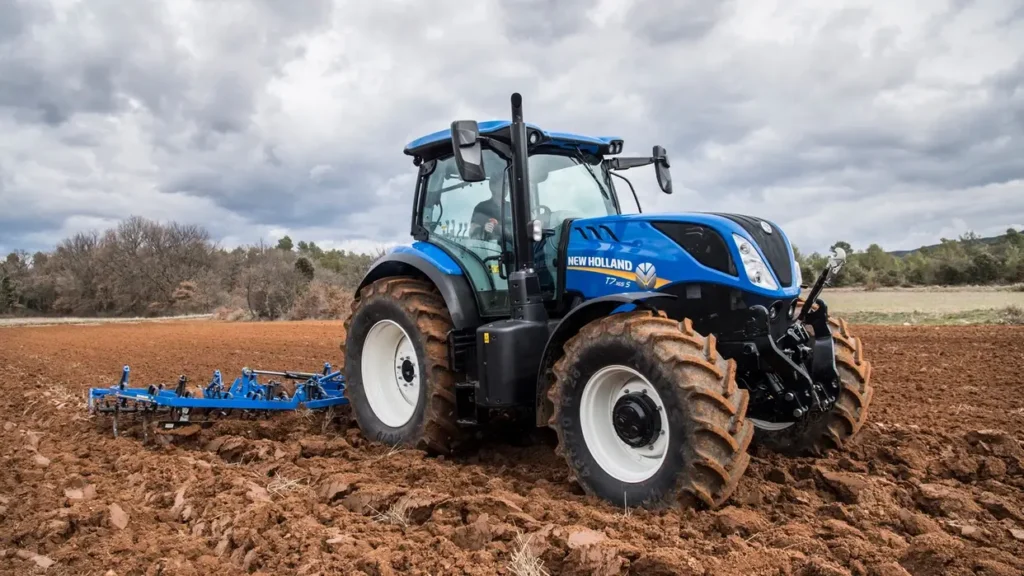 New Holland T7.265 S-traktor i arbeid på en åker, drar en harv gjennom jorden under overskyet himmel.