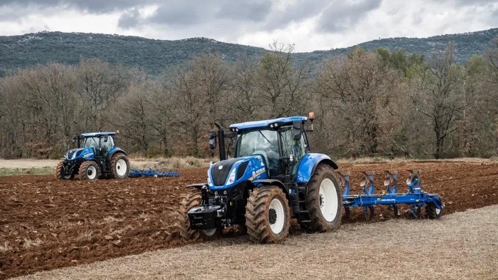 T7.265 S-traktor med plog i aksjon på en åker, med en annen traktor som arbeider i bakgrunnen. Omgitt av skog og åser.