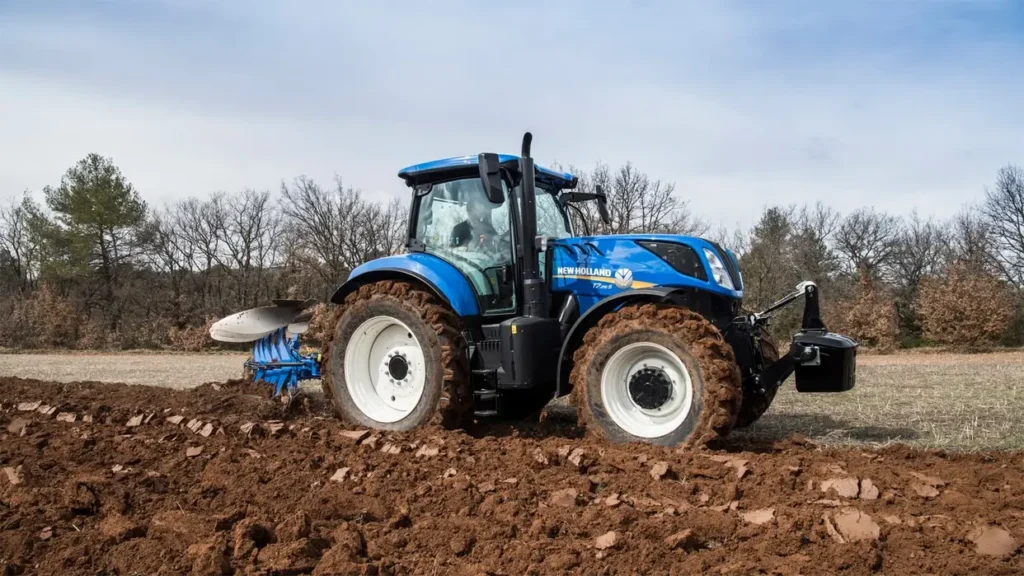 New Holland T7.265 S-traktorer pløyer en åker, omgitt av skog og fjell i bakgrunnen.