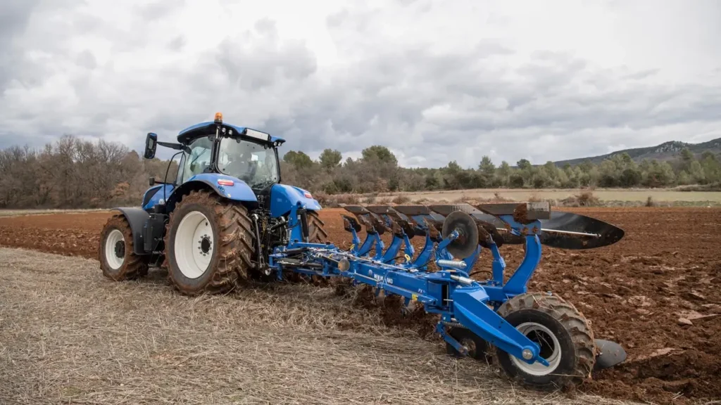 New Holland T7.265 S-traktor sett bakfra, pløyer en åker med en stor blå plog, med jordrester på skjærene.