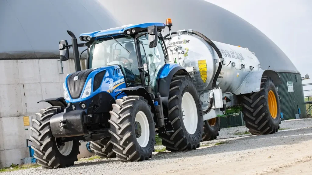 New Holland T7.265 S-traktor med en stor gjødselspreder, kjører over en gressmark med blå himmel i bakgrunnen.