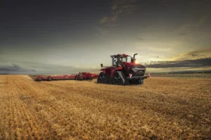 En kraftig rød Quadtrac-traktor med beltedrift jobber på en stor, gyllen åker under en dramatisk himmel i solnedgang. Traktoren er utstyrt med et stort redskap for jordbearbeiding, som strekker seg bakover, og fremhever dens kapasitet for tunge arbeidsoppgaver i landbruket.