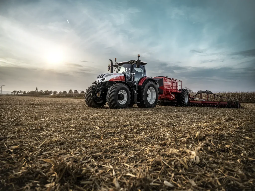 Steyr 6280 Absolut CVT traktor arbeider på et åpent jorde ved solnedgang, koblet til en rød redskap for jordbearbeiding.