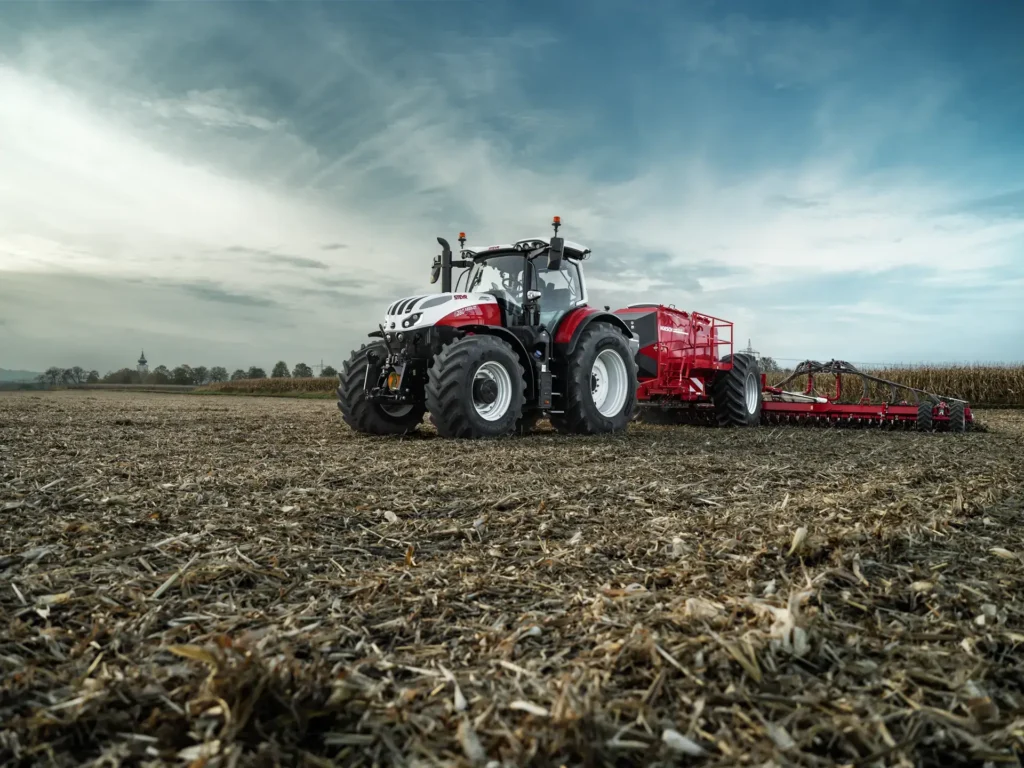 En Steyr 6280 Absolut CVT traktor trekker et rødt redskap på en pløyd åker, med desaturerte farger.