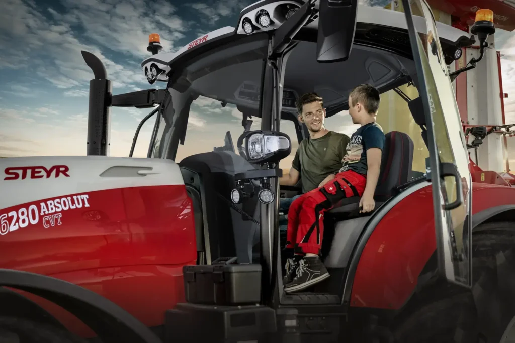 En far og sønn sitter sammen inne i førerhuset på en Steyr 6280 Absolut CVT traktor, med åpen dør.